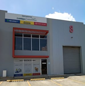 Front of a grey industrial building with a large red 8 on the right and glass storefront windows below signage panels for construction brands at the top left.