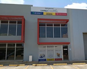 Exterior of a gray industrial building with red-framed windows and a CANELA MAQUINARIA sign above the entrance; parking lot in front.