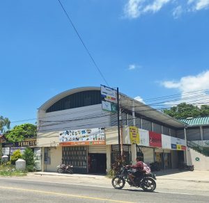 Two-story storefront on a sunny street with signs; a motorcyclist rides by in the foreground.