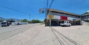 Wide street view with a two-story commercial building on the right, white pickup parked in front and clear blue sky above.